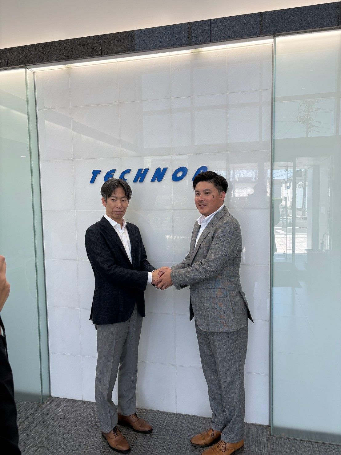 TECHNOAのロゴがある壁の前で握手を交わす、グレーのスーツ姿の若松駿太監督（右）