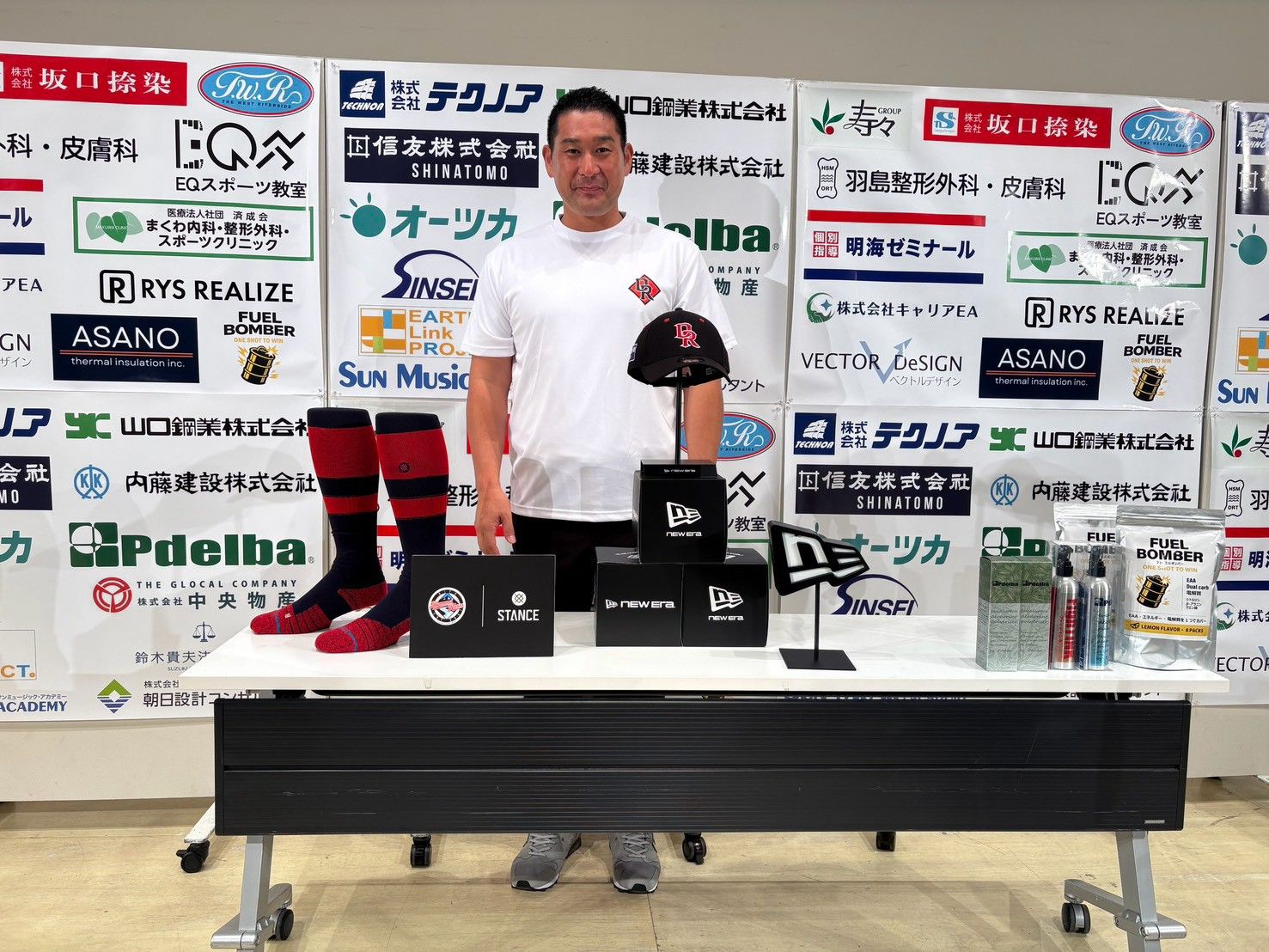 スポンサーボードの前で、NEW ERAのキャップとSTANCEのソックスを並べた机の後ろに立つ飯田祥多社長